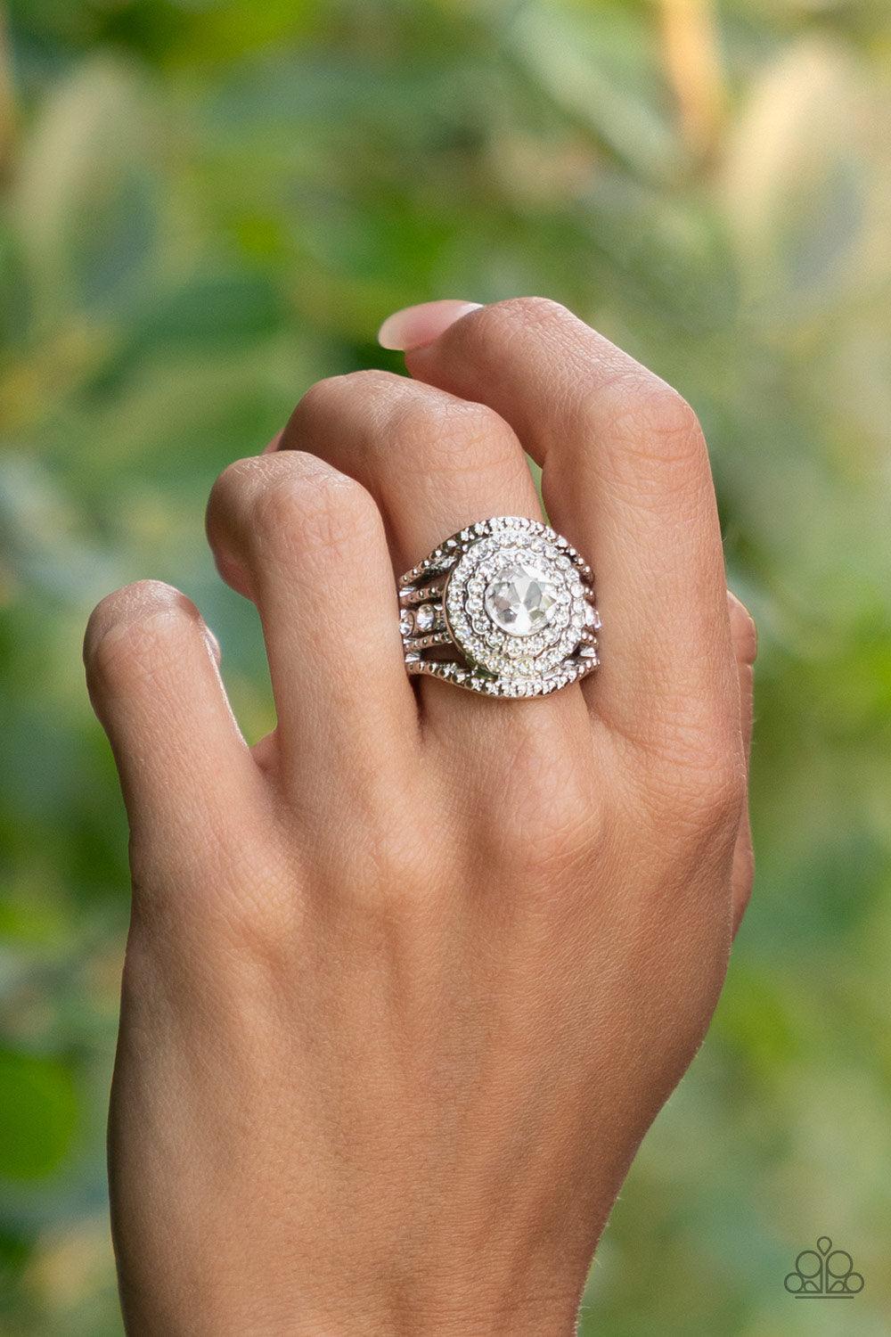 Paparazzi Accessories Understated Drama - White Bordered in stacked rings of glassy white rhinestones, a brilliant white gem sits atop layers of silver studded bars that swoop and stack into blinding bands across the finger. Features a stretchy band for a