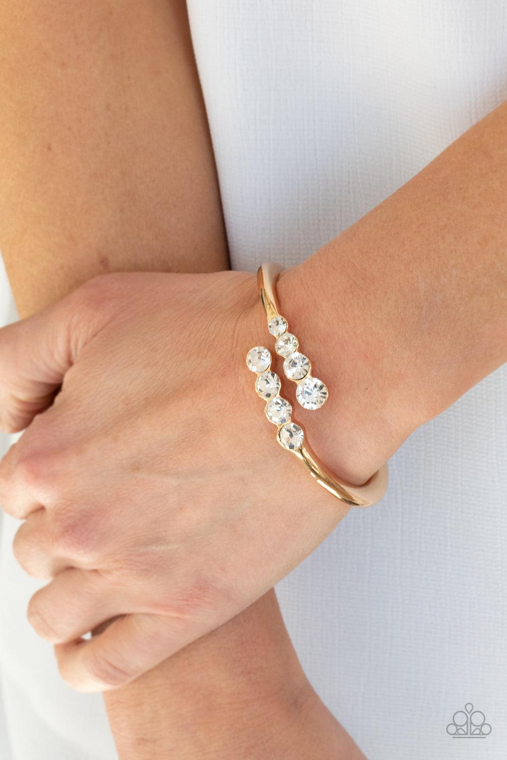 Paparazzi Accessories Defying Dazzle - Gold Attached to two substantial gold bars, oversized rows of glittery white rhinestones delicately stack into a hypnotizing centerpiece around the wrist. Features an adjustable hinge closure. Sold as one individual