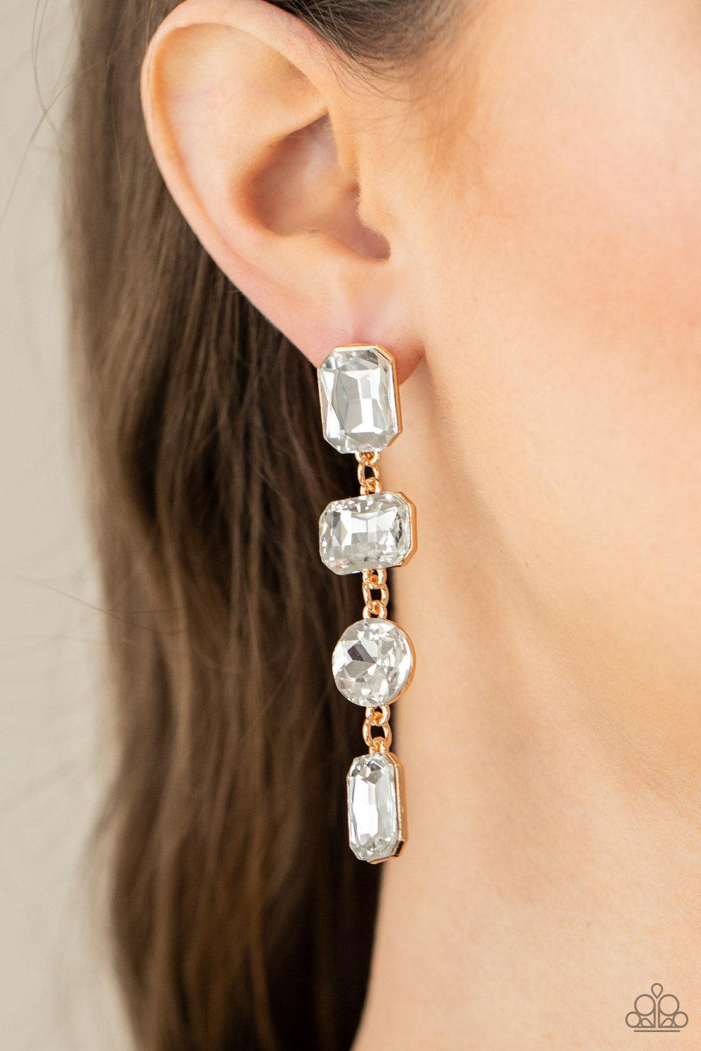 Paparazzi Accessories Cosmic Heiress - Gold A strand of oversized round, teardrop, and emerald cut white rhinestones trickles from the ear, creating a jaw-dropping golden chandelier. Earring attaches to a standard post earring. Sold as one pair of post ea