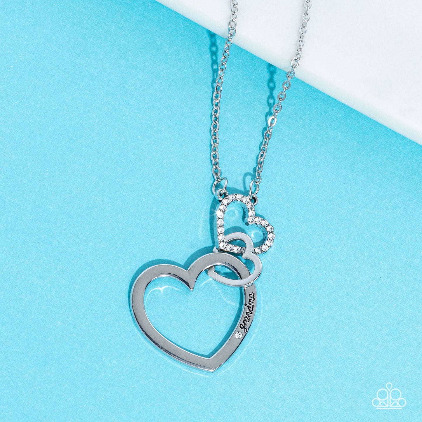 Paparazzi Accessories Grandma Glow - White A silver heart engraved with the word "Grandma," dangles from two smaller hearts at the bottom of a lengthened silver chain. The first heart is accented with sparkly white rhinestones creating an affectionately s