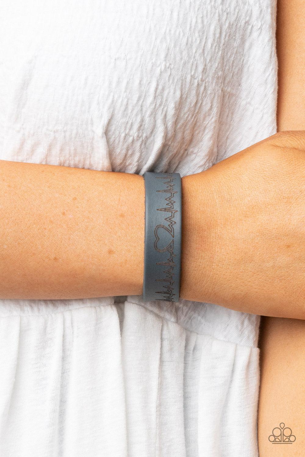 Paparazzi Accessories Haute Heartbeat - Silver Culminating into a heart at the center, a zigzagging pattern reminiscent of a heartbeat is etched across the front of a gray leather band around the wrist for a whimsical fashion. Features an adjustable snap