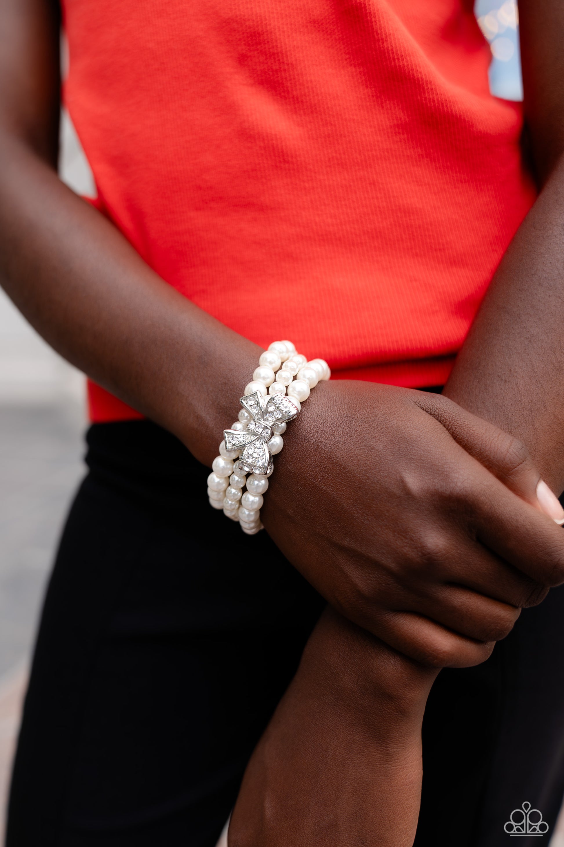Paparazzi Accessories How Do You Do? - White Strung along elastic stretchy bands, three strands of high-sheen classic white pearls wrap around the wrist. Featured atop the pearly collection, a loopy silver bow, filled with glistening white rhinestones is