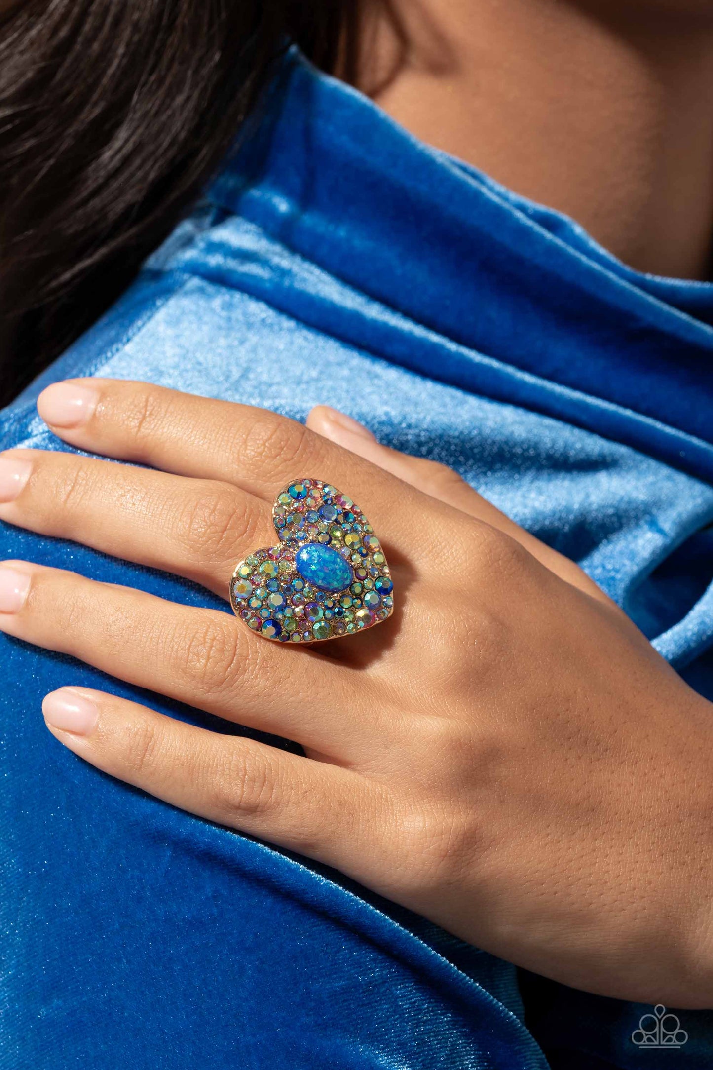 Paparazzi Accessories Bejeweled Beau - Blue Encrusted in brilliant multicolored and iridescent rhinestones, a glittery gold heart frame adorns the center of an airy gold band for a flirtatious finish. Featuring iridescent flecks, an opalescent blue oval b