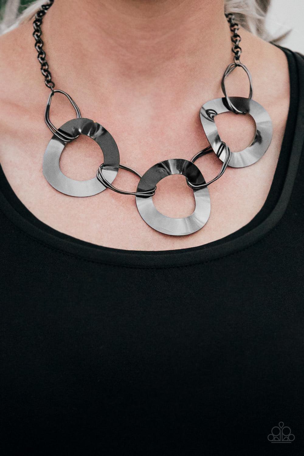 Paparazzi Accessories Modern Mechanics - Black Featuring slightly warped surfaces, asymmetrical gunmetal hoops link with reflective gunmetal rings below the collar for a modern look. Features an adjustable clasp closure. Sold as one individual necklace. I