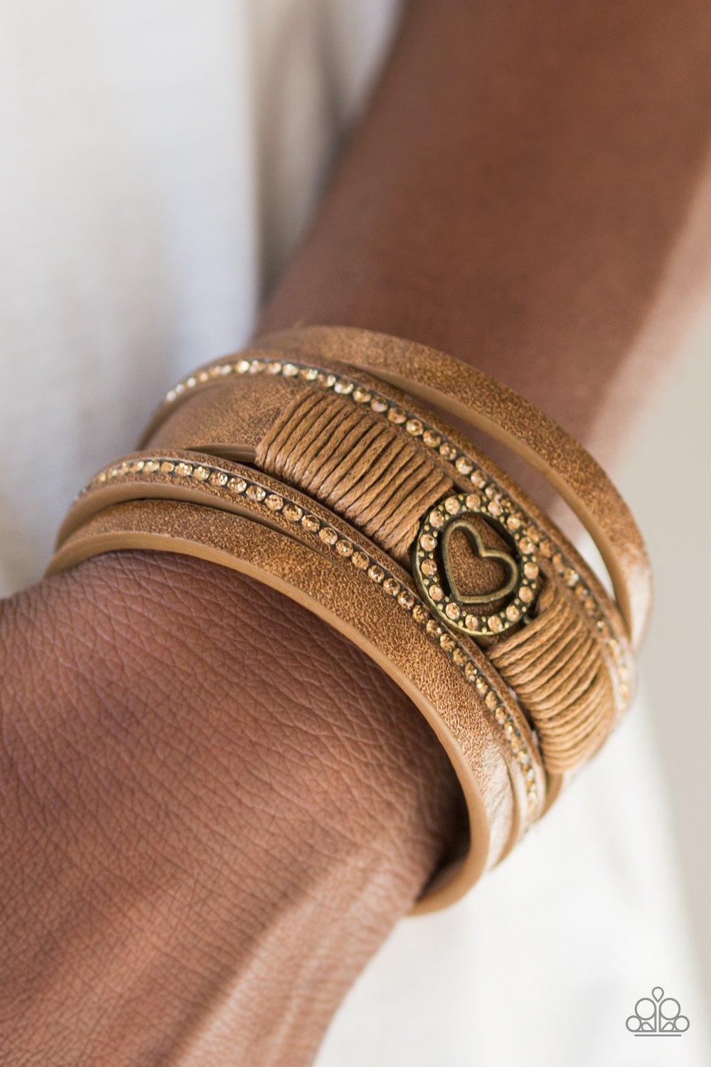 Paparazzi Accessories It Takes Heart - Brass Brushed in a metallic shimmer, strips of brown leather are encrusted in alternating rows of glittery topaz rhinestones. Brown cording knots around the centermost strand, securing a sparkling heart frame in plac