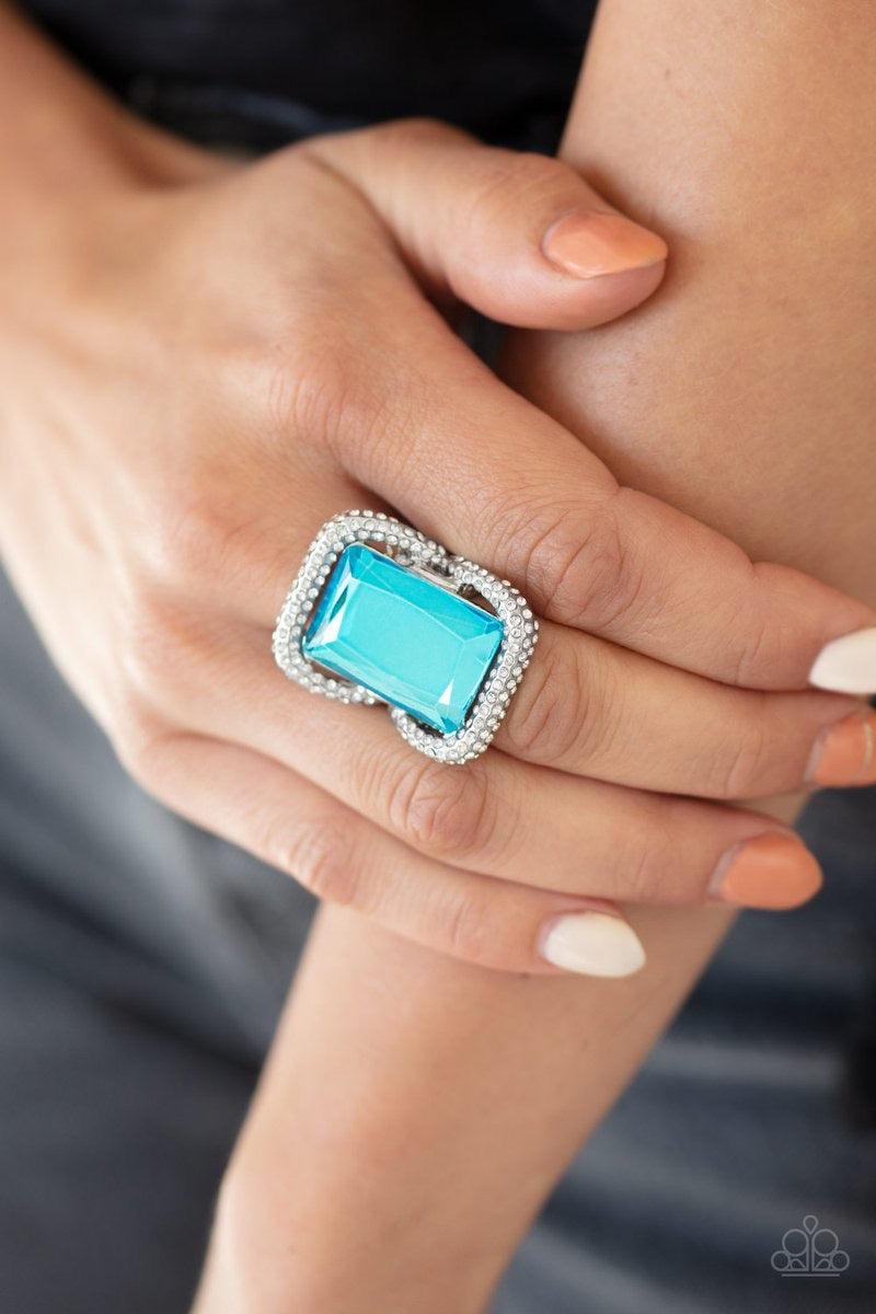 Paparazzi Accessories Deluxe Decadence - Blue Featuring a regal emerald style cut, an oversized blue gem nestles inside a rounded square silver frame encrusted in glassy white rhinestones for a dramatically glamorous look. Features a stretchy band for a f