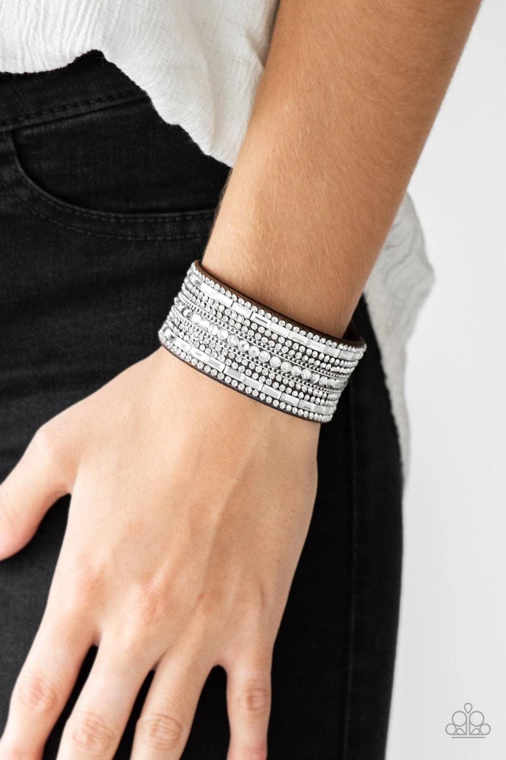 Paparazzi Accessories Rebel Radiance - Brown Featuring classic round and edgy emerald style cuts, glittery white rhinestones and glistening silver chains are encrusted along bands of brown suede for a sassy look. Features an adjustable snap closure. Sold