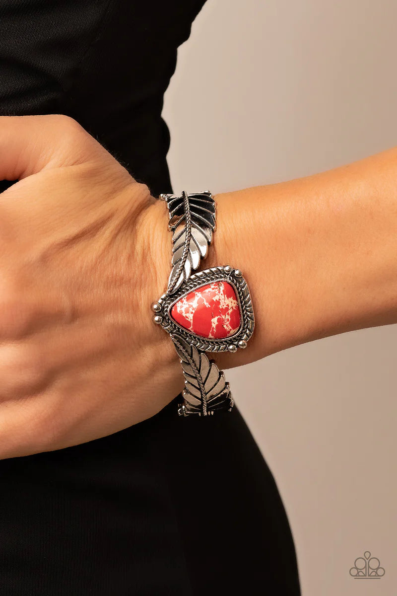 Paparazzi Accessories Desert Roost - Red A twisted metal bar lines the center of an elongated silver feather as it curves into a rustic cuff. Featuring a studded and textured silver frame, an asymmetrical red stone is balanced between the ends of the silv