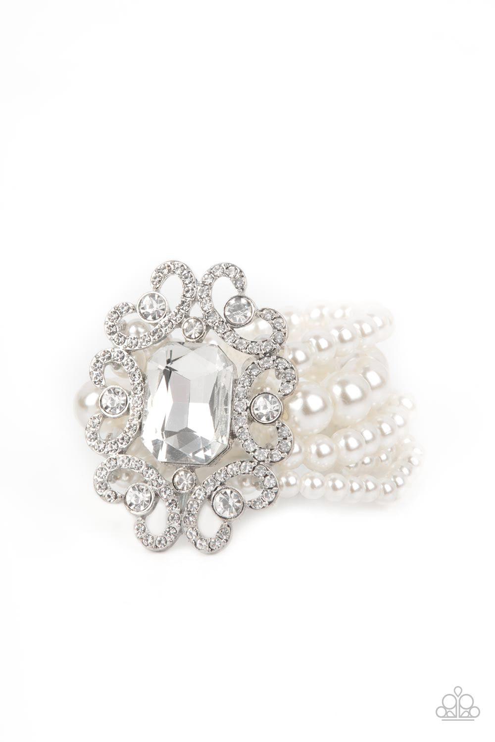 Paparazzi Accessories Rule The Room - White A dramatically oversized emerald cut gem adorns the center of a white rhinestone encrusted filigree filled frame. The blinding centerpiece attaches to bubbly rows of pearl beaded stretchy bracelets, creating a g