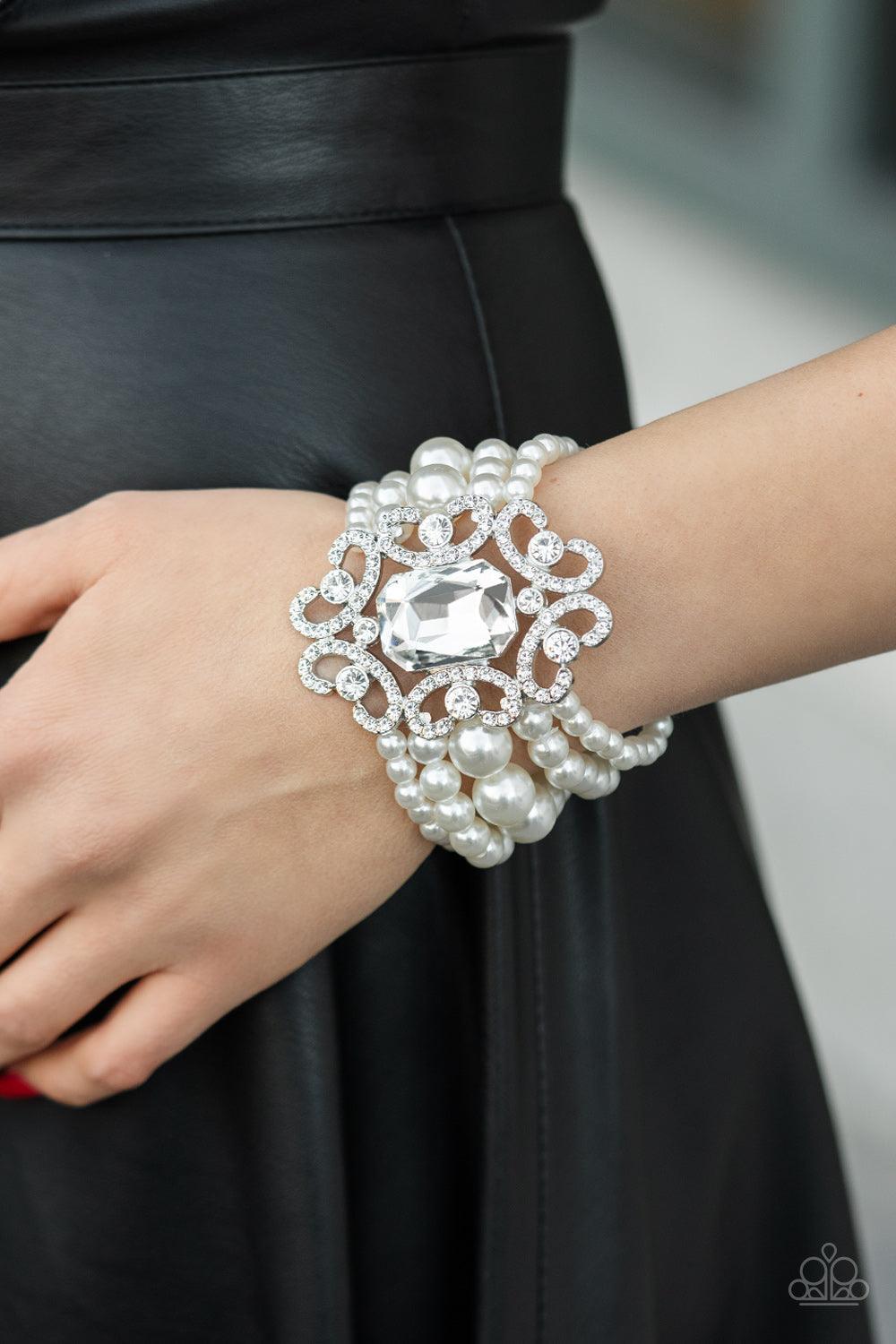 Paparazzi Accessories Rule The Room - White A dramatically oversized emerald cut gem adorns the center of a white rhinestone encrusted filigree filled frame. The blinding centerpiece attaches to bubbly rows of pearl beaded stretchy bracelets, creating a g