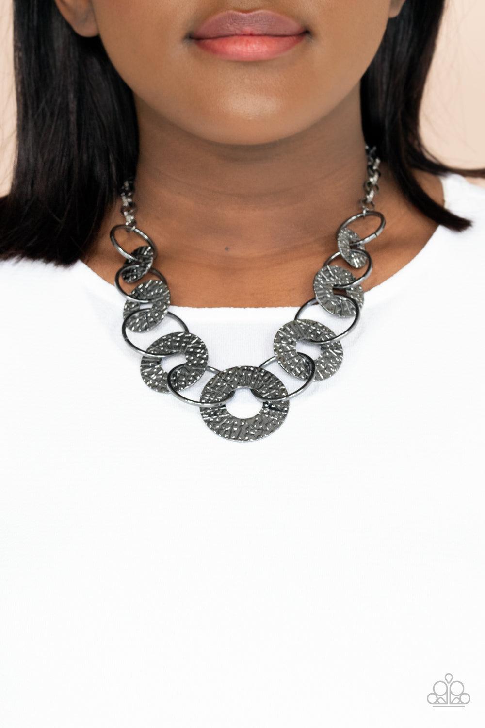 Paparazzi Accessories Industrial Envy - Black Gradually increasing in size near the center, a collection of glistening gunmetal rings and abstract hammered discs interlock below the collar for an intense industrial look. Features an adjustable clasp closu
