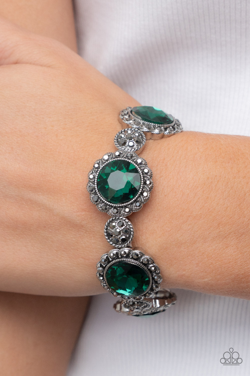 Paparazzi Accessories Palace Property - Green An oversized green rhinestone adorns the center of a hematite rhinestone petaled floral frame. Infused with hematite dotted silver accents, the glitzy floral frames sparkle along stretchy bands around the wris