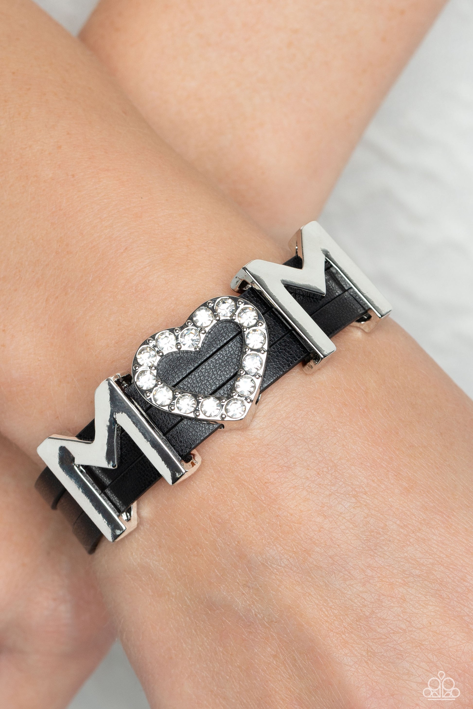 Paparazzi Accessories Heart of Mom - Black A row of glassy white rhinestones are encased into a sparkly heart frame that is threaded along layers of black leather strands around the wrist. Framing both sides of the heart frame, two high-sheen silver lette