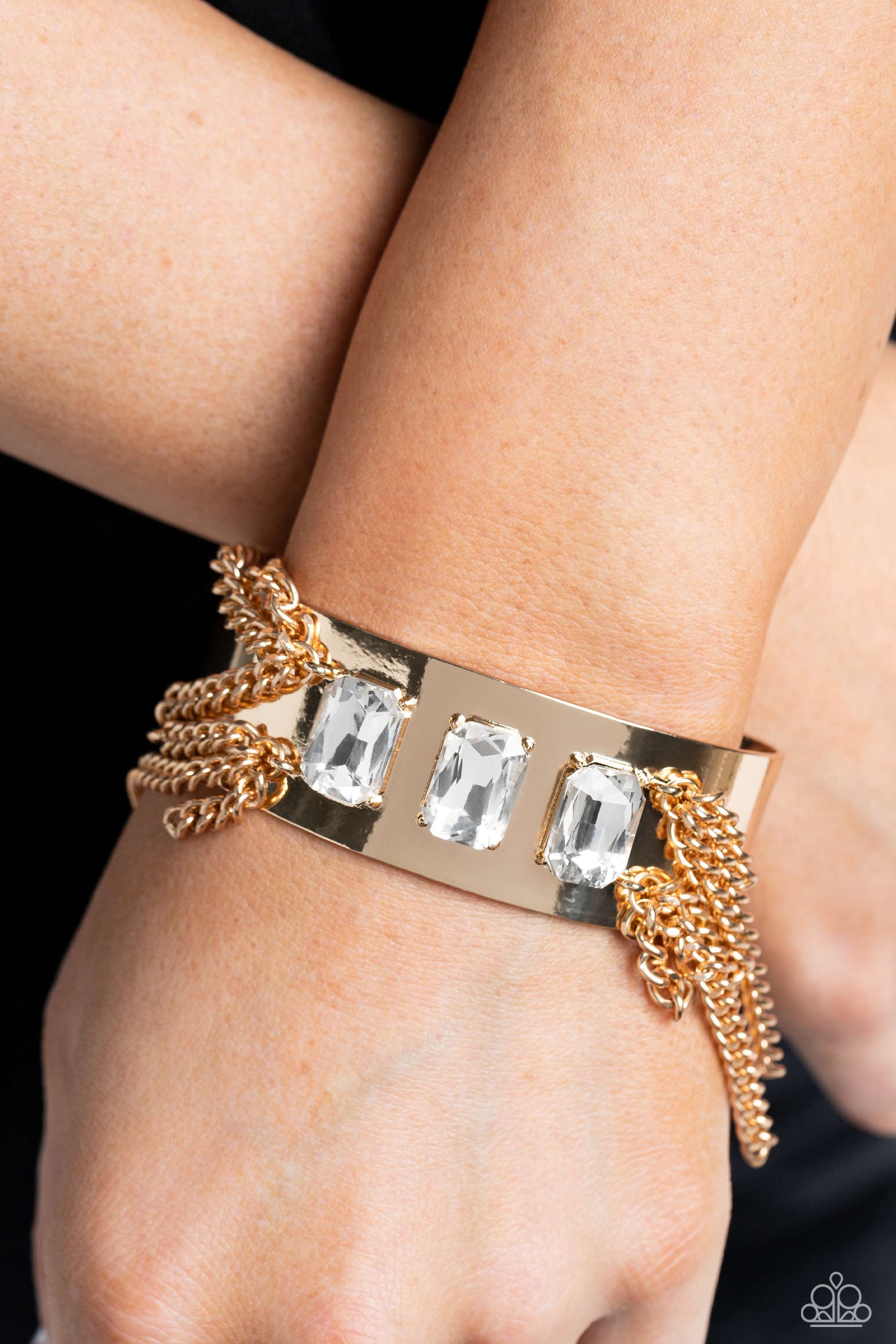 Paparazzi Accessories CHAIN Showers - Gold A thick, industrial gold cuff is topped by three brilliant white, emerald-cut gems. Strands of gold curb chain stream from each side of the trio of glitz, adding undeniable drama and eye-catching movement to the
