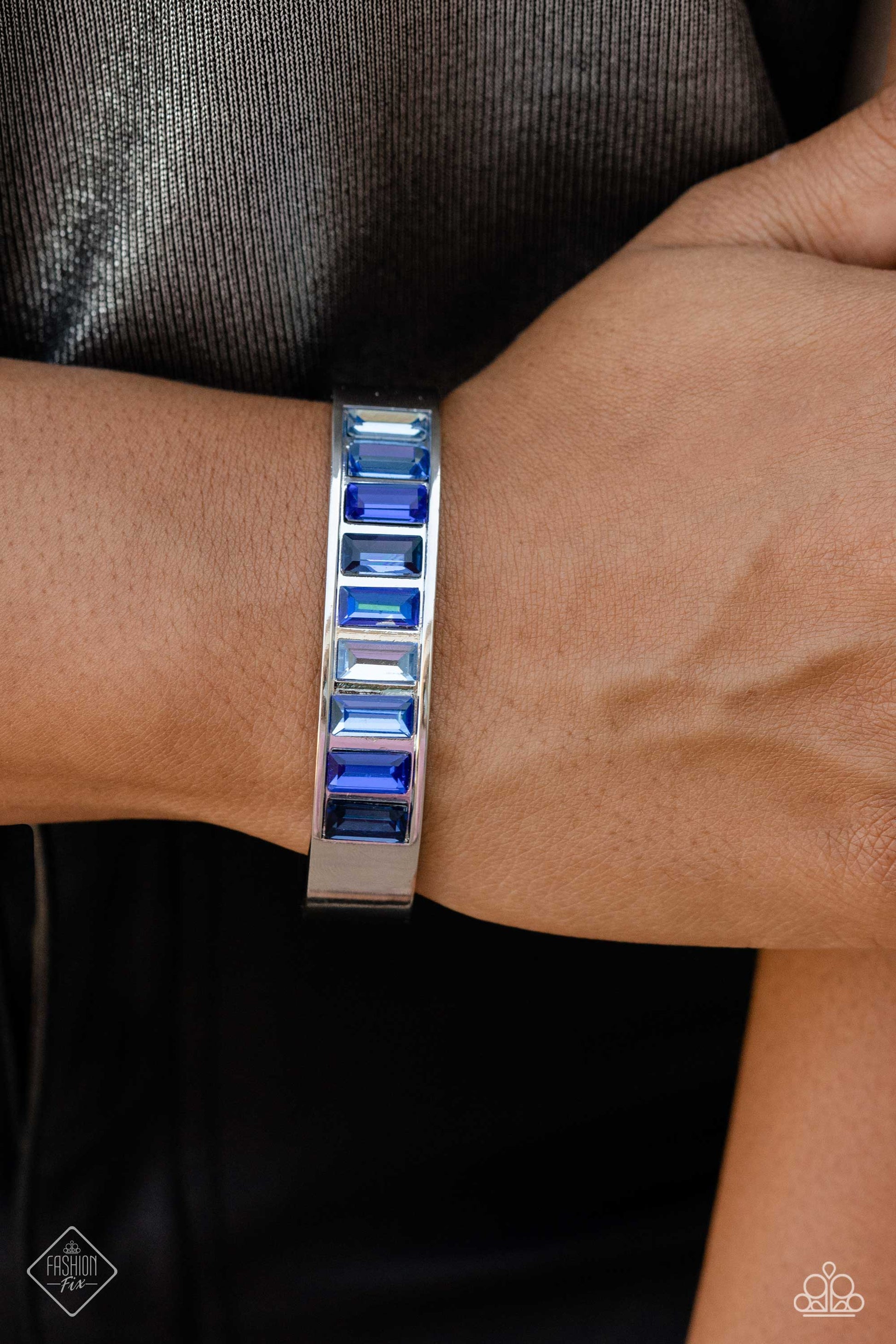 Paparazzi Accessories Practiced Poise - Blue Embedded in the center of a thick, high-sheen silver cuff, a row of glassy emerald-cut gems in varying shades of blue radiates atop the wrist, creating an unexpected pop of color. Sold as one individual bracele