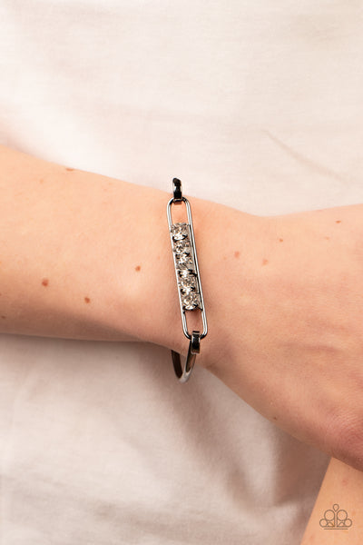 Paparazzi Accessories In CHARM’S Way - Black Encased in gunmetal square fittings, a row of glassy white rhinestones are fitted in place along an airy gunmetal frame that hinges to the center of a bangle-like bracelet for a timeless finish. Features a hing