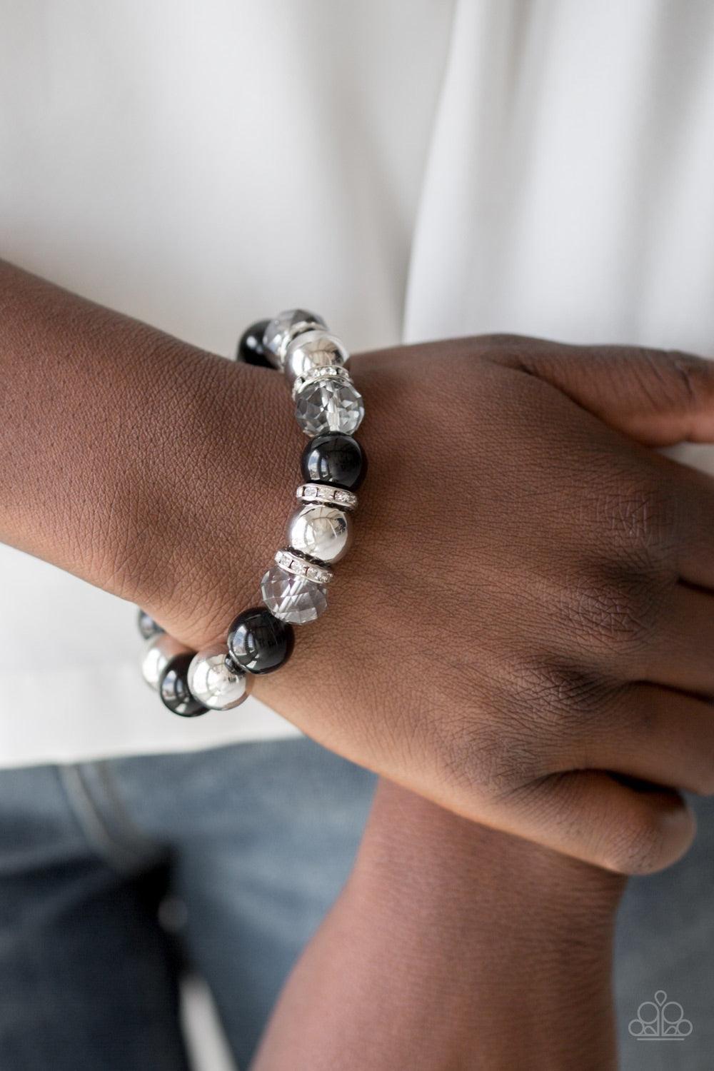 Paparazzi Accessories Camera Chic - Black Oversized silver, smoky crystal-like, and polished black beads are threaded along a stretchy elastic band and wrapped around the wrist for a glamorous look. White rhinestone encrusted rings are sprinkled between t