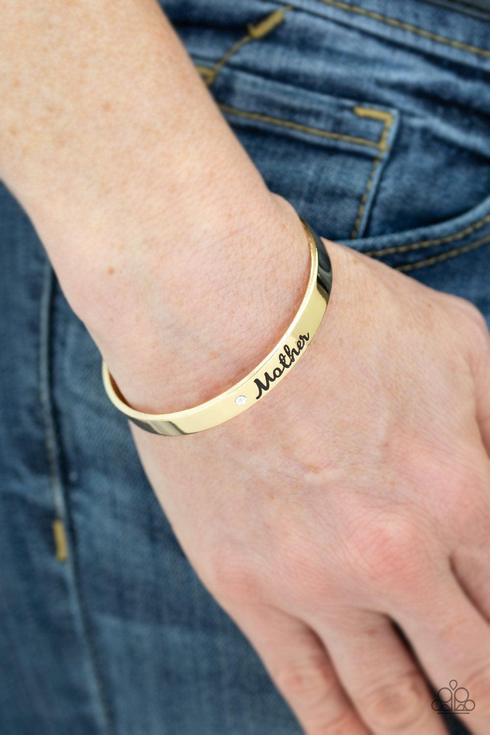 Paparazzi Accessories Every Day Is Mother’s Day - Gold A sleek gold cuff wraps around the wrist in a casual fashion. Dotted with a dainty white rhinestone, the shimmery cuff is engraved in the word “Mother” for a loving finish. Jewelry