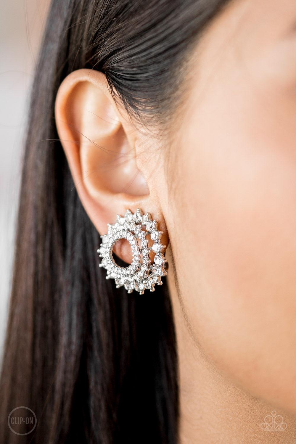 Paparazzi Accessories Buckingham Beauty - White *Clip-On Gradually increasing in size, glassy white rhinestones are encrusted along three stacking silver frames for a regal look. Earring attaches to a standard clip-on fitting. Sold as one pair of clip-on