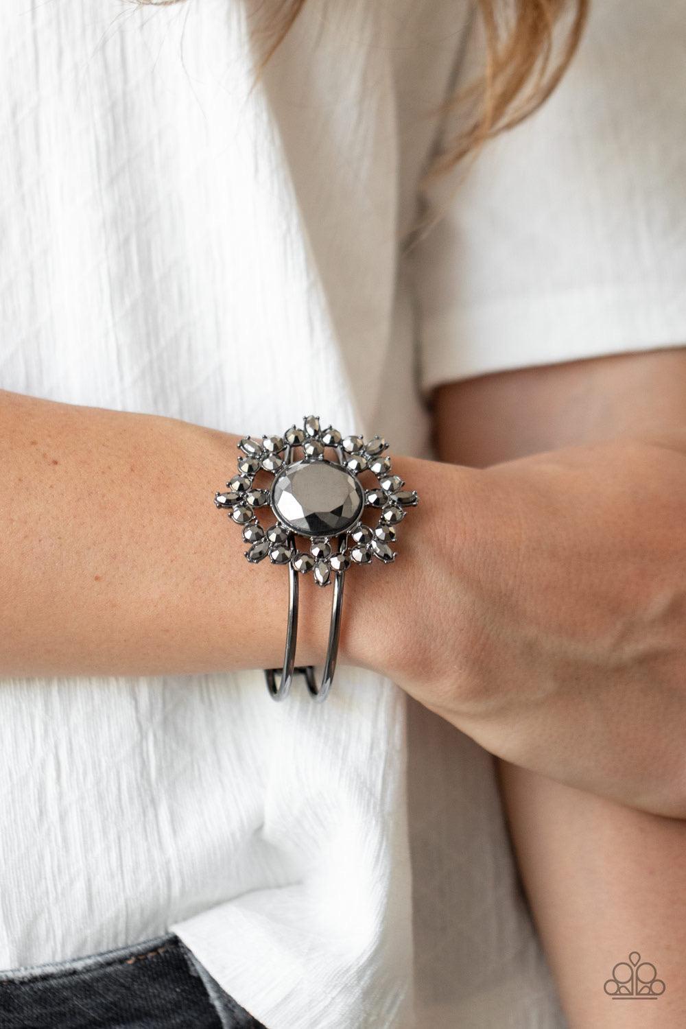 Paparazzi Accessories Elaborate Elegance - Black Glittery hematite petals bloom from an oversized hematite rhinestone center, creating a dazzling floral centerpiece atop an airy gunmetal cuff. Features a hinged closure. Sold as one individual bracelet. Je