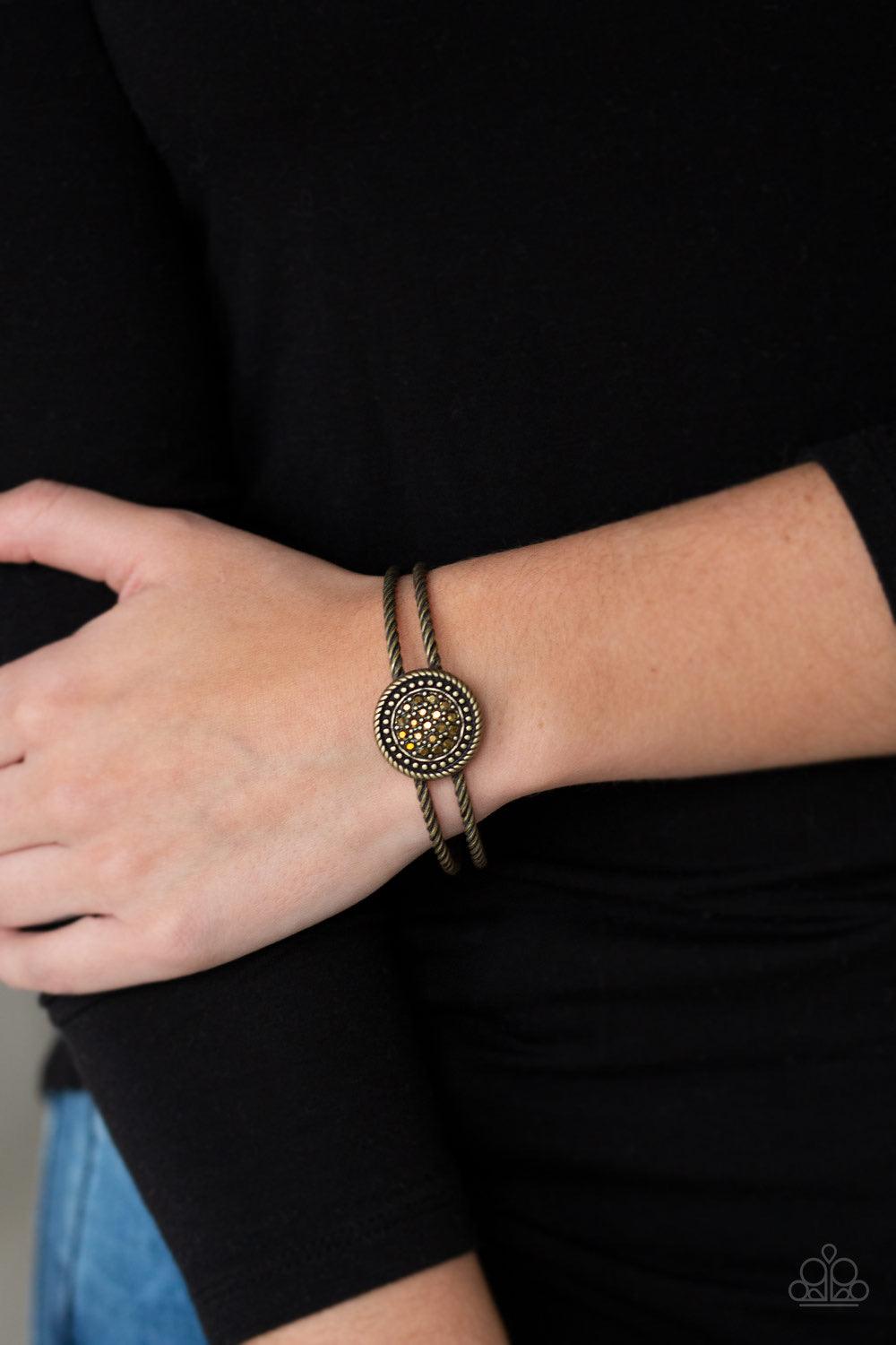 Paparazzi Accessories Definitely Dazzling - Brass Brushed in an antiqued shimmer, twisted brass bars arc across the finger, coalescing into an airy cuff. Radiating in brass studs and glittery aurum rhinestones, a round brass frame adorns the center for an