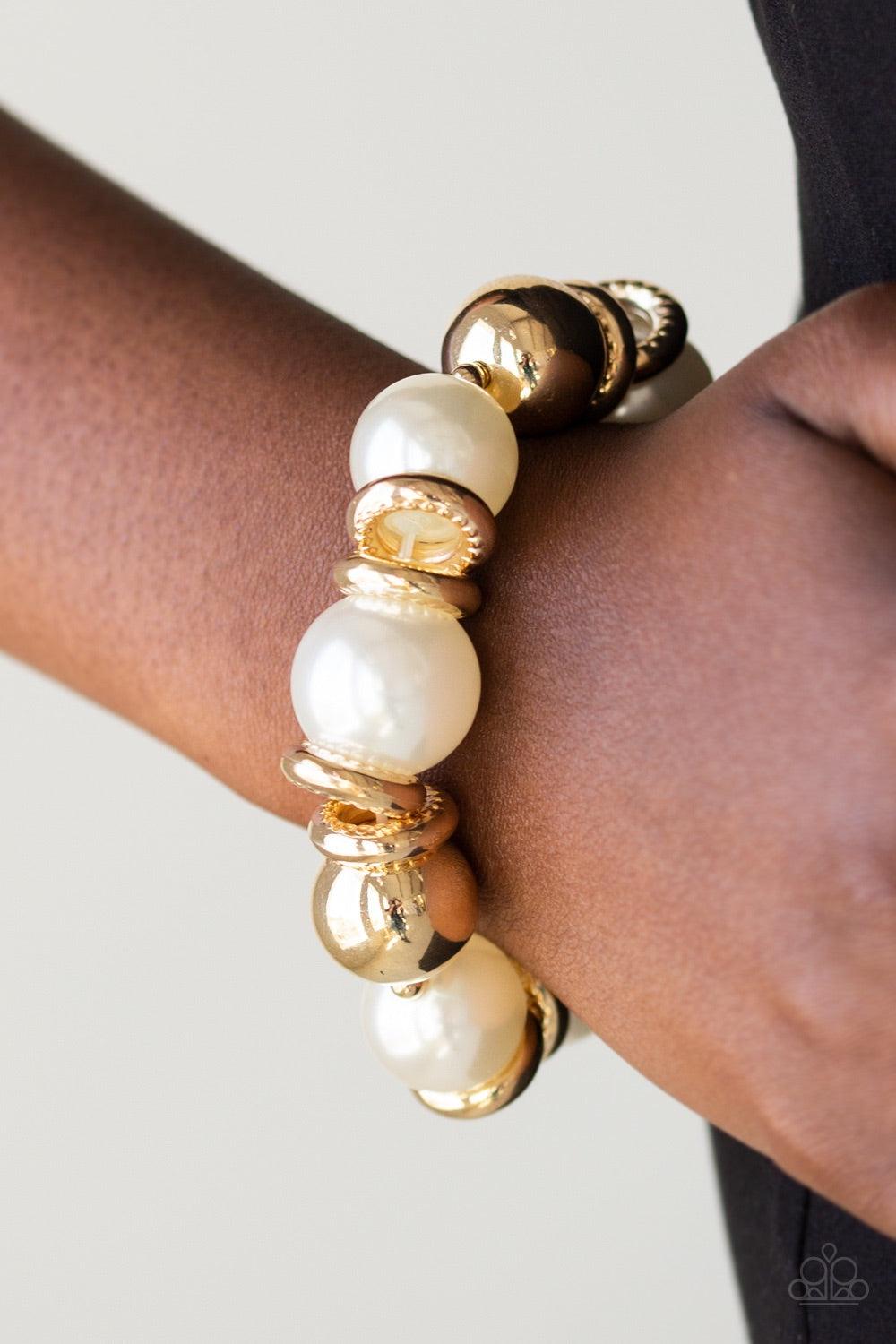 Paparazzi Accessories Rockin Rockefeller - Gold Dramatic white pearls and shiny gold beads are threaded along an elastic stretchy band around the wrist. Glistening gold accents are sprinkled between the classic beads, adding depth and shimmer to the bubbl
