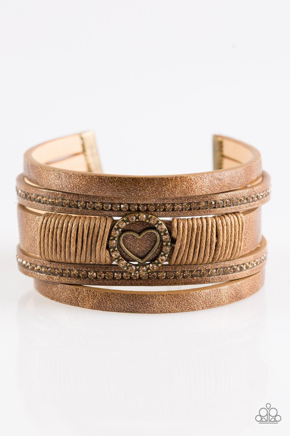Paparazzi Accessories It Takes Heart - Brass Brushed in a metallic shimmer, strips of brown leather are encrusted in alternating rows of glittery topaz rhinestones. Brown cording knots around the centermost strand, securing a sparkling heart frame in plac