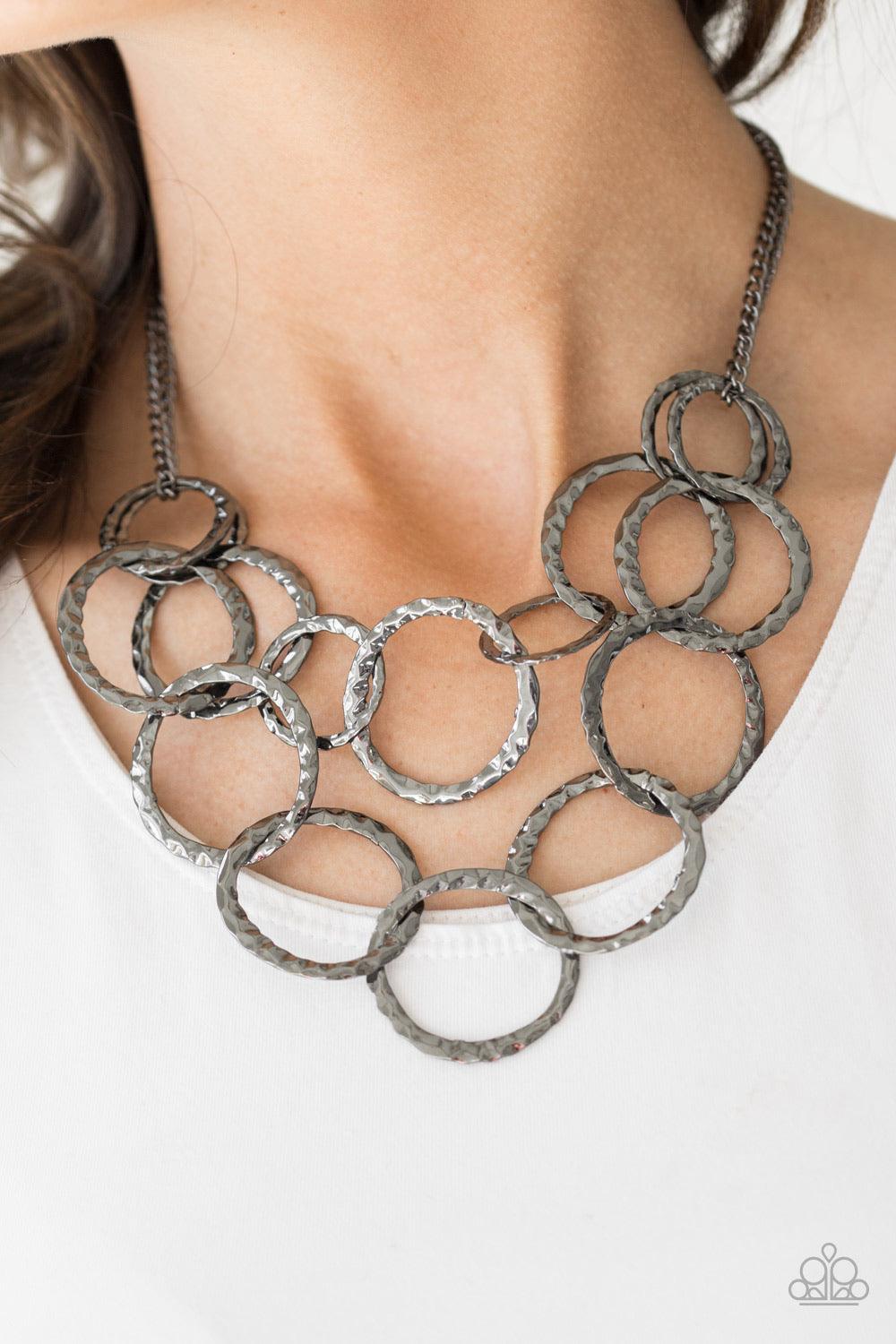 Paparazzi Accessories Radiant Ringmaster - Black Varying in size, gunmetal rings are delicately hammered with light catching shimmer. The glistening rings link below the collar, draping into two layered rows for an edgy finish. Features an adjustable clas
