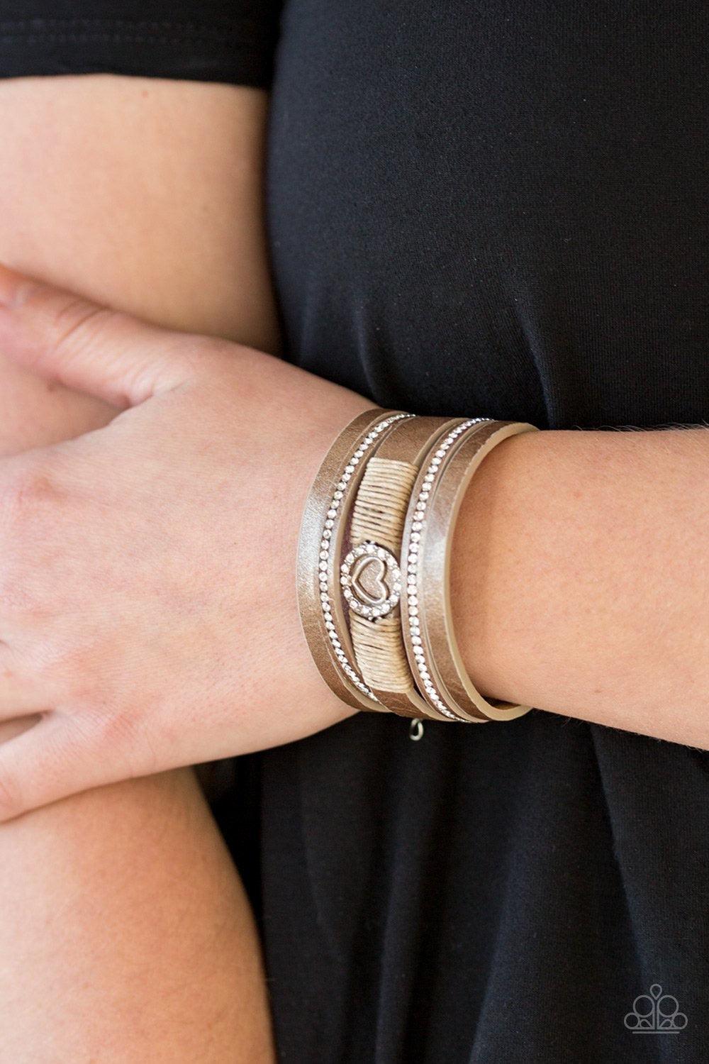 Paparazzi Accessories It Takes Heart - Brown Brushed in a metallic shimmer, strips of brown leather are encrusted in alternating rows of glittery white rhinestones. Brown cording knots around the centermost strand, securing a sparkling heart frame in plac
