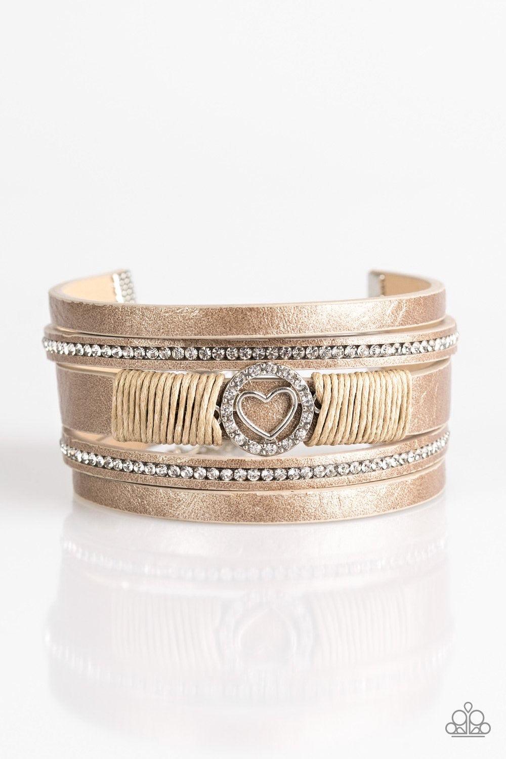 Paparazzi Accessories It Takes Heart - Brown Brushed in a metallic shimmer, strips of brown leather are encrusted in alternating rows of glittery white rhinestones. Brown cording knots around the centermost strand, securing a sparkling heart frame in plac
