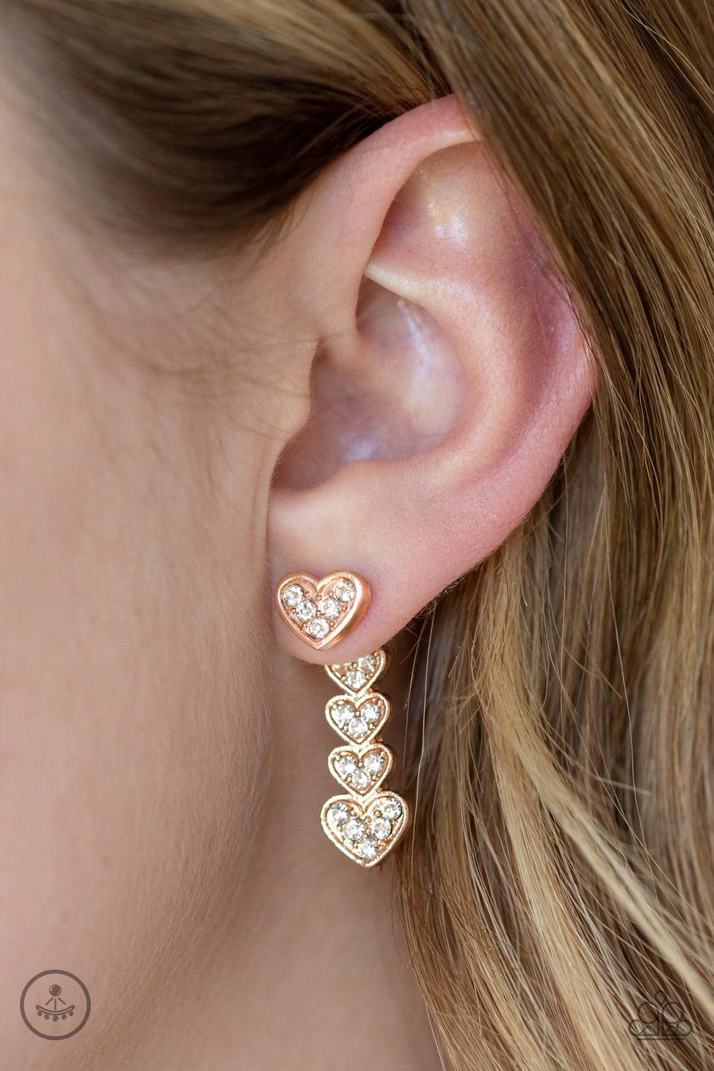 Paparazzi Accessories Heartthrob Twinkle - Rose Gold A white rhinestone encrusted rose gold heart attaches to a double-sided post, designed to fasten behind the ear. Stacked in gradually increasing white rhinestone encrusted heart frames, the glittery dou