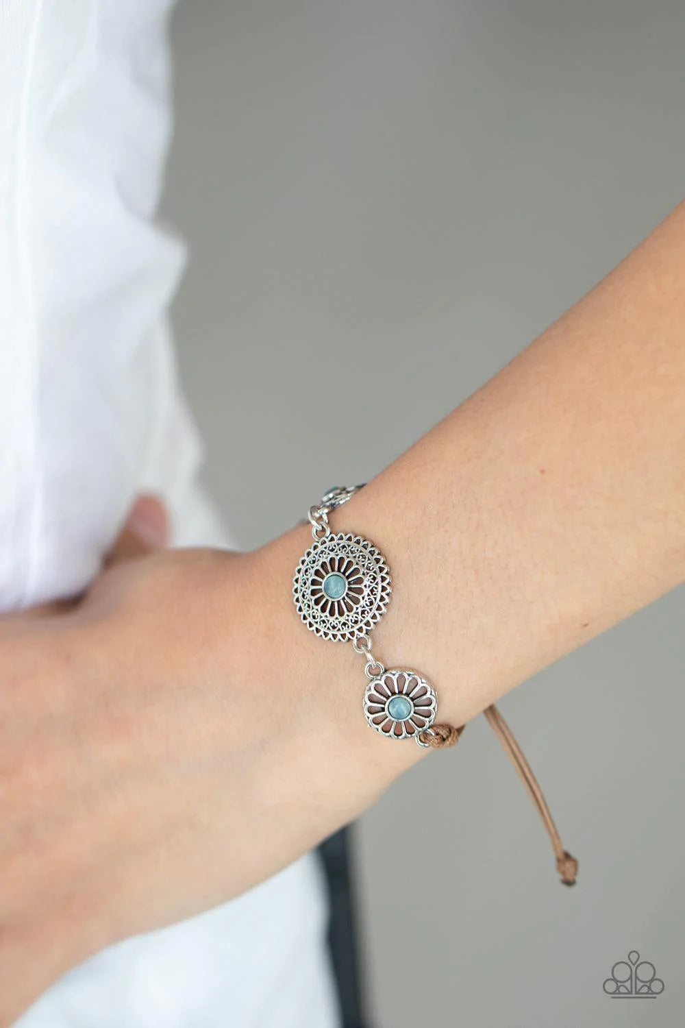 Paparazzi Accessories Bohemian Botany - Blue Dotted with dainty blue cat's eye stone centers, an airy trio of ornate floral frames delicately link across the wrist. The whimsy centerpiece is knotted in place around the wrist with shiny brown cording for a