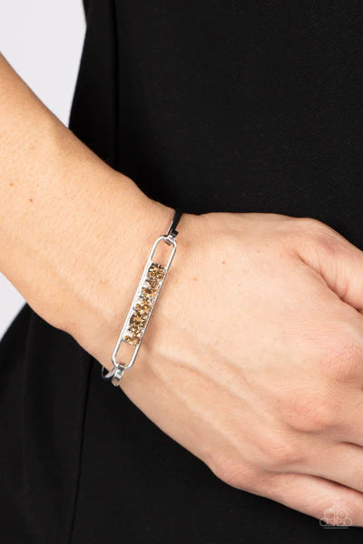 Paparazzi Accessories In CHARM’S Way - Brown Encased in silver square fittings, a row of golden topaz rhinestones are fitted in place along an airy silver frame that hinges to the center of a bangle-like bracelet for a timeless finish. Features a hinged c